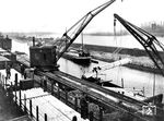 Güterumschlag im Rheinhafen Duisburg-Hochfeld mittels dampfbetriebenen Breitspurkran von den Rungenwagen der Reichsbahn auf einen Frachtkahn. Am jenseitigen Rheinufer ist die Silhouette des Krupp-Hüttenwerks Rheinhausen zu erkennen. (1930) <i>Foto: RVM</i>