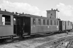 Zu den KB4trp 970-877, KB4trp 970-878, KB4trp 970-885 und KB4tr 970-890 des Prignitzer Netzes: Von der Waggon- und Maschinenfabrik AG (Bautzen) und der Sächsischen Waggonfabrik Werdau erwarben die K.Sächs.Sts.E.B. in den Jahren 1913/14 insgesamt 95 Traglastenwagen (Gattung 729) mit der 1913 eingeführten 4. Klasse sowie einem genieteten Untergestell aus Stahlprofilen mit Sprengwerk. Nach dem Krieg folgten 144 weitere Wagen. Gegenüber der Gattung 720 waren sie um 78 cm kürzer, aber 18 cm breiter. Neben einem Großraum mit 20 Doppel-Querbänken boten sie ein Traglastenabteil mit zwei Längsbänken sowie einen Abort ohne Waschgelegenheit. Die offenen Perrons hatten wiederum offene Übergangseinrichtungen. Nach dem Entfall der 4. (1928) und der 3. Klasse (1956) boten die Wagen nur noch die 2. Klasse. Anfangs besaßen sie innen lediglich zwei Lampen, mit den Jahren kamen weitere hinzu. Die sieben schmalen Fenster je Längsseite in unterschiedlichen Abständen waren herablaßbar. Über ihnen erstreckten sich über die gesamte Wagenlänge hölzerne Gepäckablagen. Ende der 1940er Jahre gab die RBD Dresden vier solcher Wagen ans Perleberger Netz ab, und zwar je zwei Bautzener und Werdauer. Von jedem Fabrikat wurde ein Exemplar in der Prignitz etwa Mitte der 1950er Jahre außen verblecht, während die Wagen 970-878 und -890 bis zuletzt ihre Holzbeplankung behielten. Hier ist der Wagen 970-877 im Sommer 1963 in Glöwen in einen sächsischen Zug eingereiht, der von einer Österreicherin – der 99 4712 – gezogen wird.  (1963) <i>Foto: Birger Wilcke (Sammlung Ludger Kenning)</i>